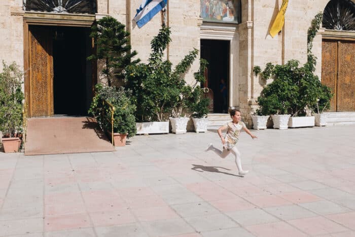 A Young girl runnign through a square with the Greek Flag behind her.